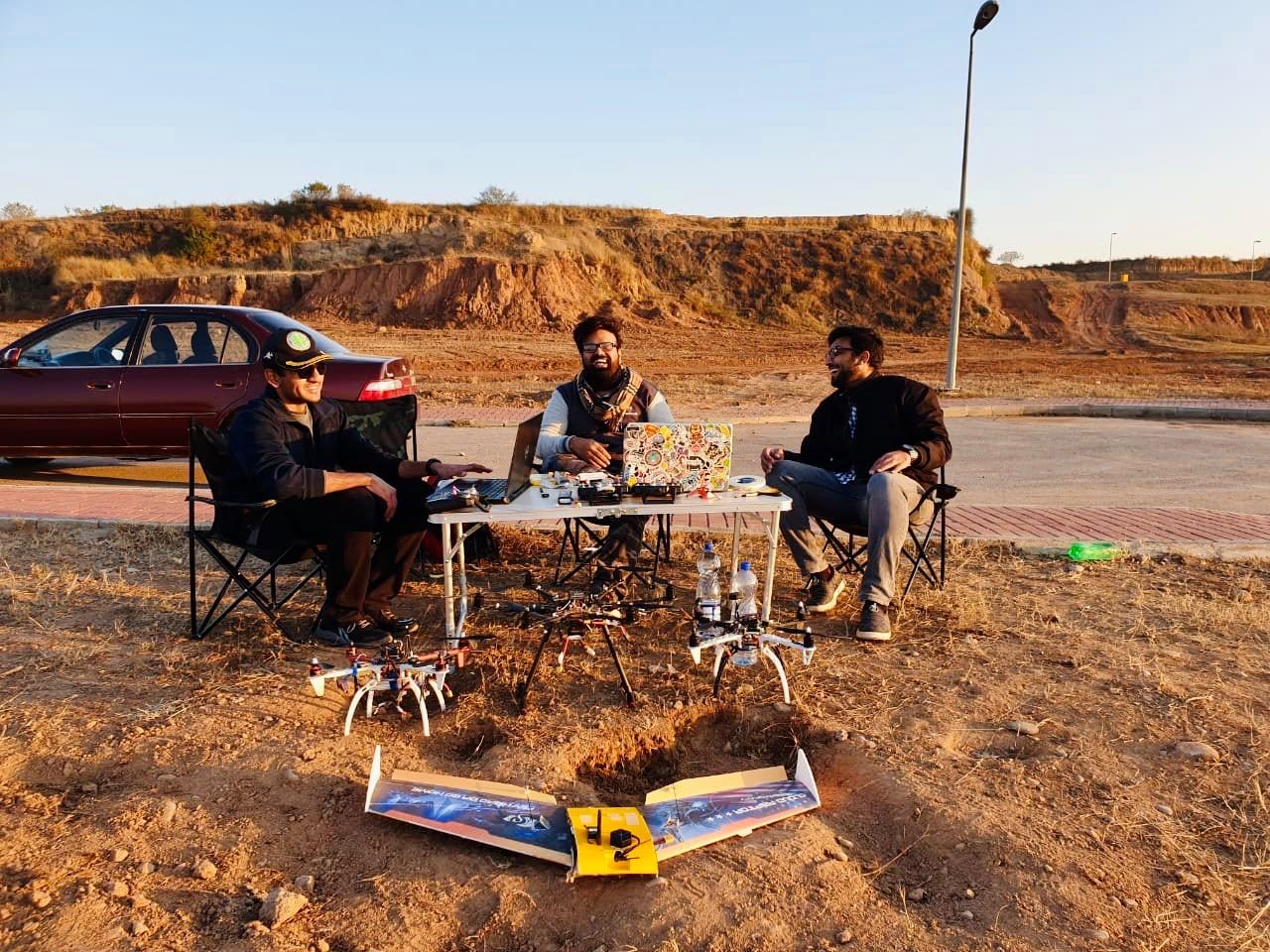 Field team seated at a folding table on a dirt embankment beside a fixed-wing UAV.